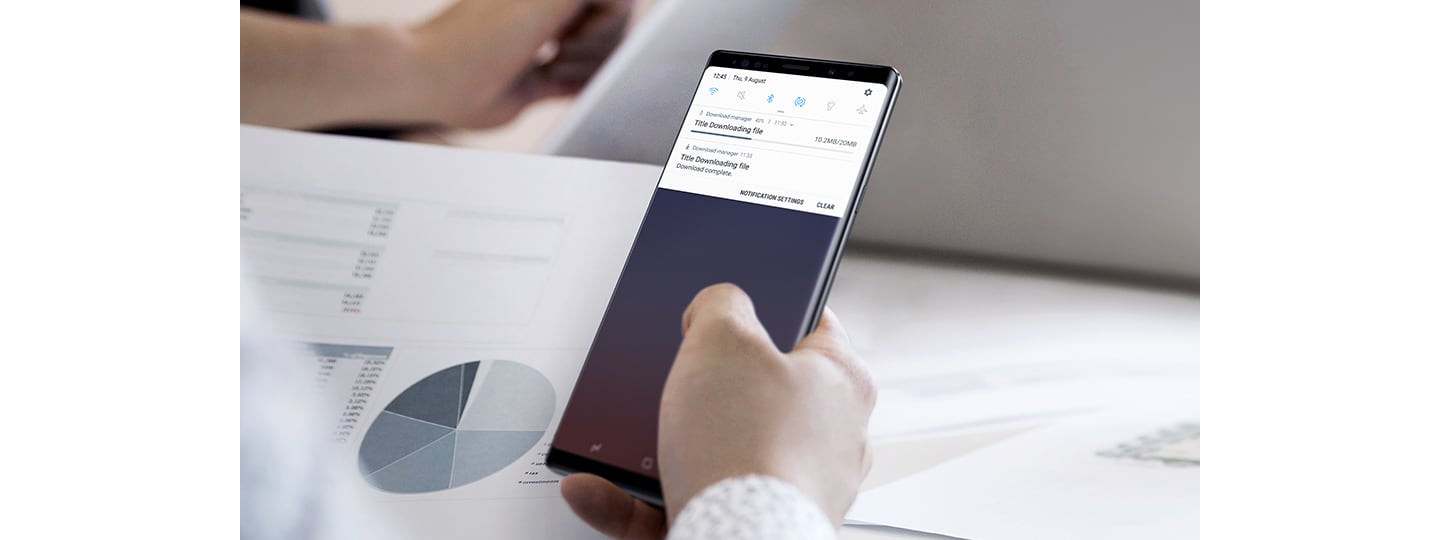 Vaizdas su žmogumi, laikančiu „Galaxy Note9“, o ekrane rodomas atsisiuntimo ekrano vaizdas, vienas atsisiuntimas beveik baigtas, o kitas – užbaigtas