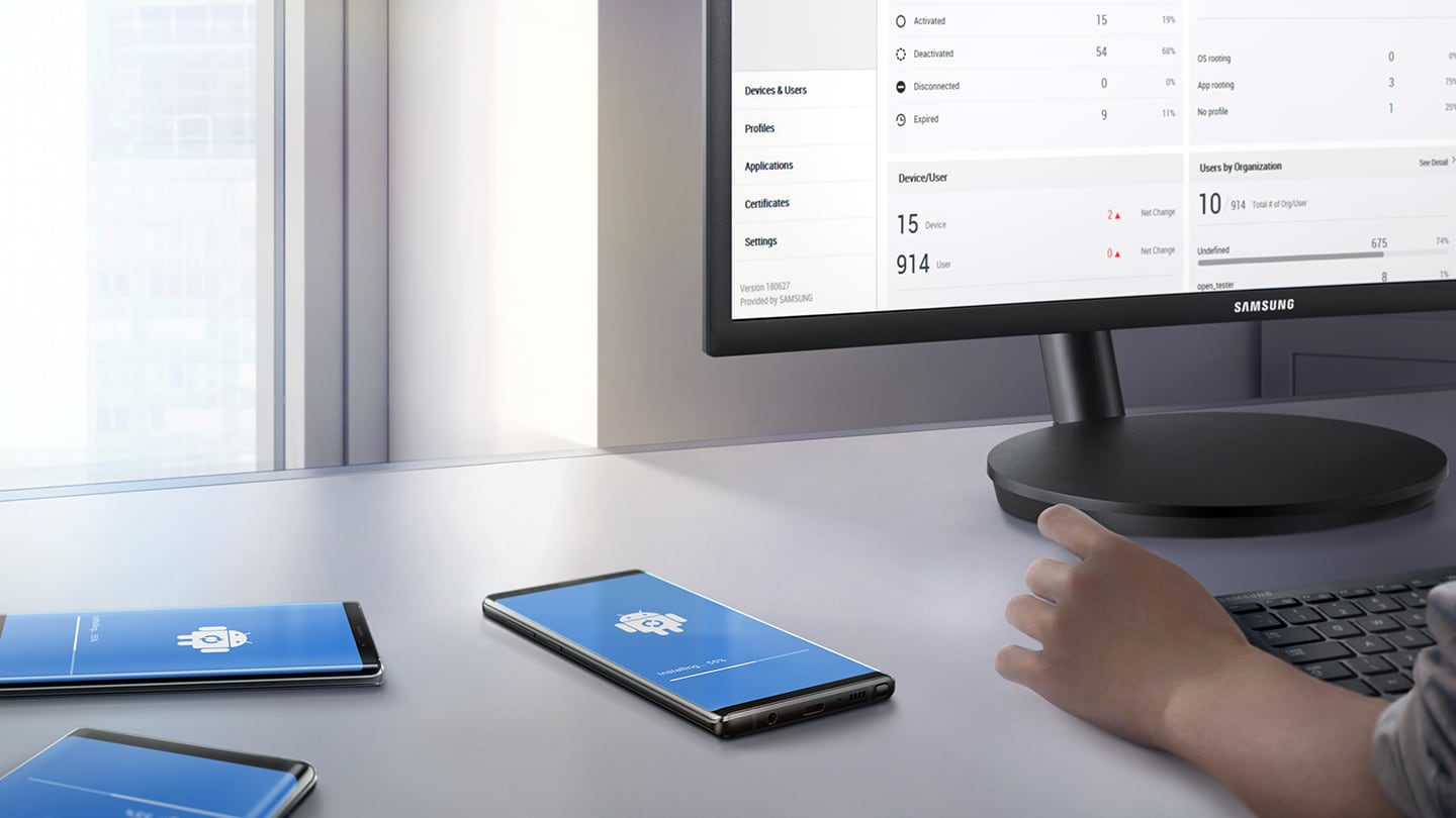 Vaizdas su žmogumi prie stalo su keliais „Galaxy Note9“, kurių ekrane matomas „Android“ atnaujinimo vaizdas ir „Knox Manage“ vaizdas monitoriuje