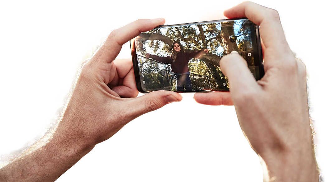 Image of a person taking a photo with Galaxy S9+ with a photo displayed on-screen and the scene shown in the background to demonstrate OIS
