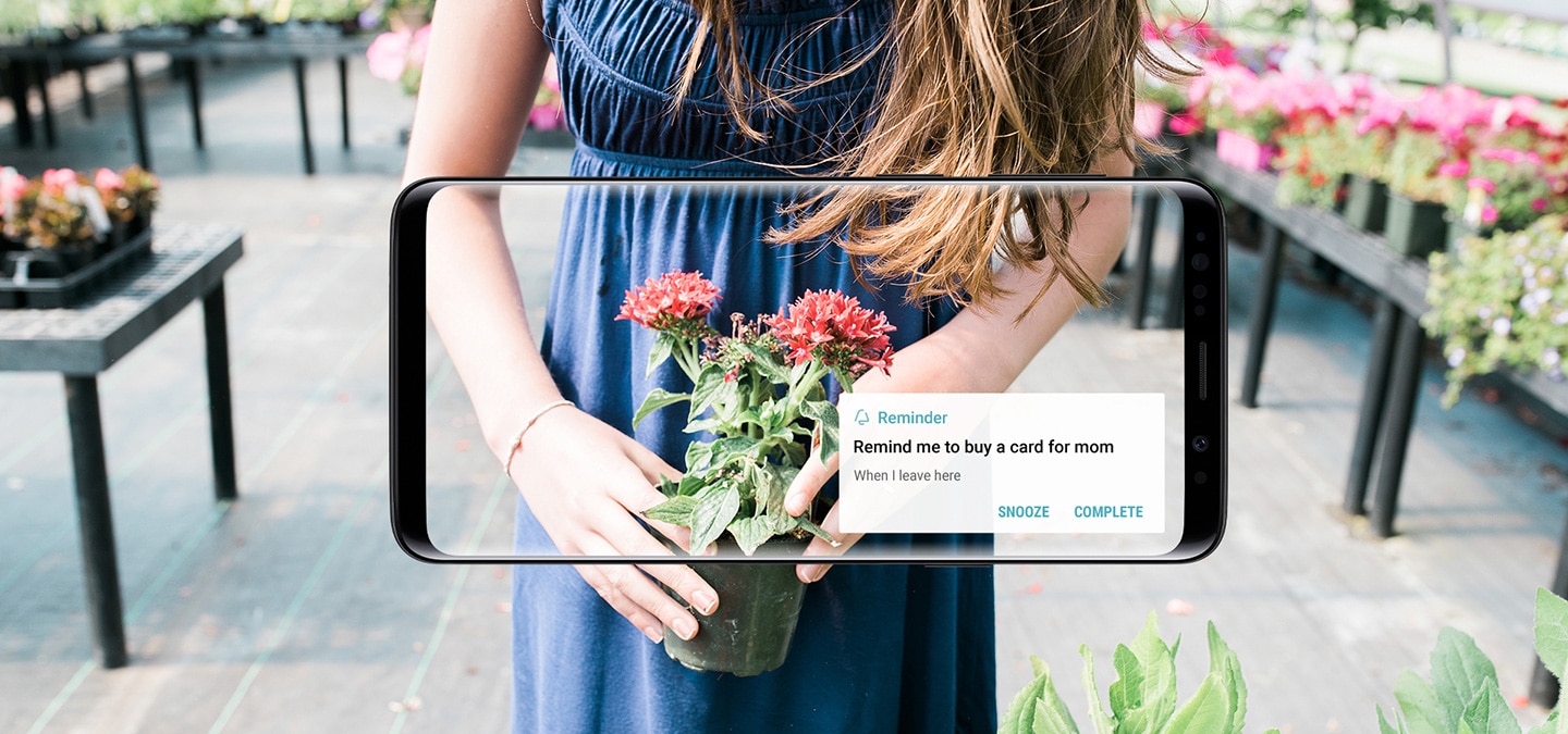 Вид спереди смартфона Galaxy S9 Midnight Black в интерьере цветочного магазина; на экране показано уведомление “Перед уходом не забыть купить открытку для мамы”, отравленное приложением “Напоминания Bixby”.