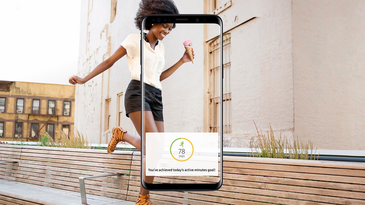 Jeune femme marchant dans le parc et application Samsung Health indiquant « Vous avez établi aujourd'hui un nouveau record du nombre de pas. » Vue avant d'un Galaxy S9 Midnight Black recevant une notification