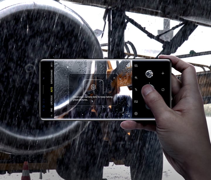 Imagem da pessoa a segurar o Galaxy Note9 e a tirar uma foto a um motor de avião à chuva, com “Limpe a lente da câmara para tirar ótimas fotos” no ecrã, demonstrando a Deteção de Falhas