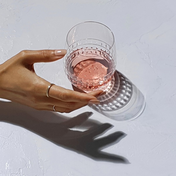 A photo taken by Galaxy Note9 of a cut crystal glass containing a pink liquid, and a hand extended to grab the glass, all casting shadows on a white surface