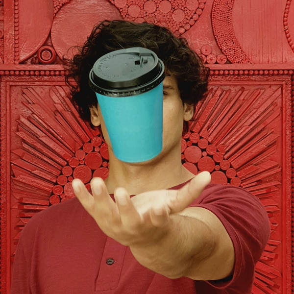 A photo taken by Galaxy Note9 of a man wearing a red shirt, standing against a red wall, throwing a blue paper cup with black lid into the air