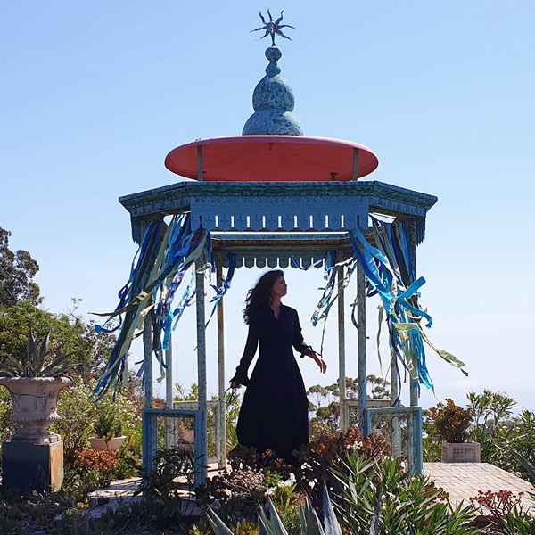 A photo taken by Galaxy Note9 of a woman wearing a long dress, standing in a blue and red gazebo surrounded by greenery